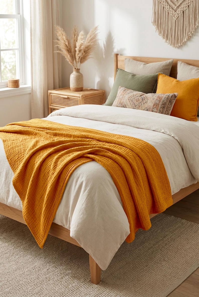 K&ouml;nnte beinhalten: Eine senfgelbe, texturierte Baumwoll-Musselin-Tagesdecke, die &uuml;ber einem Bett mit wei&szlig;em Bettbezug liegt, mit einem Holzbettrahmen, einem Nachttisch aus Rattan und dekorativen Kissen in Olivgr&uuml;n, Senfgelb und einem gemusterten neutralen Farbton.