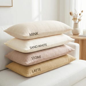 Pode incluir: Quatro almofadas retangulares em tons de bege, creme, rosa e castanho empilhadas em um sof&aacute; branco. As almofadas s&atilde;o rotuladas como "Mink", "Sand White", "Stone" e "Latte". As almofadas t&ecirc;m uma superf&iacute;cie texturizada.