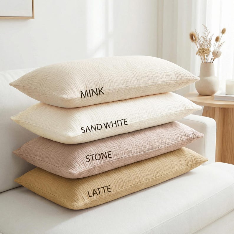 Pode incluir: Quatro almofadas retangulares em tons de bege, creme, rosa e castanho empilhadas em um sof&aacute; branco. As almofadas s&atilde;o rotuladas como "Mink", "Sand White", "Stone" e "Latte". As almofadas t&ecirc;m uma superf&iacute;cie texturizada.