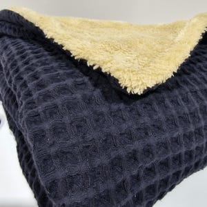 May include: A folded navy blue waffle-weave blanket with a soft, cream-coloured sherpa lining. The blanket is folded to display both textures and colours, offering a cosy and inviting aesthetic. The waffle weave provides a textured surface.