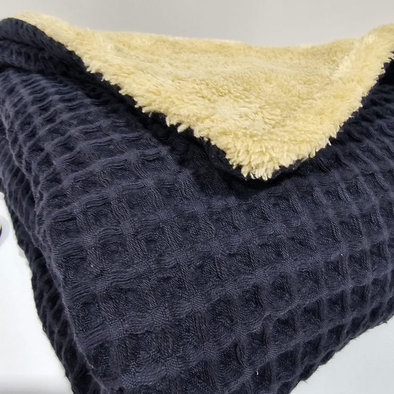 May include: A folded navy blue waffle-weave blanket with a soft, cream-coloured sherpa lining. The blanket is folded to display both textures and colours, offering a cosy and inviting aesthetic. The waffle weave provides a textured surface.