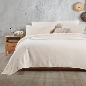 May include: A white waffle-weave throw blanket on a bed with white pillows. The bed is in a room with a white wall and a wooden bedside table.