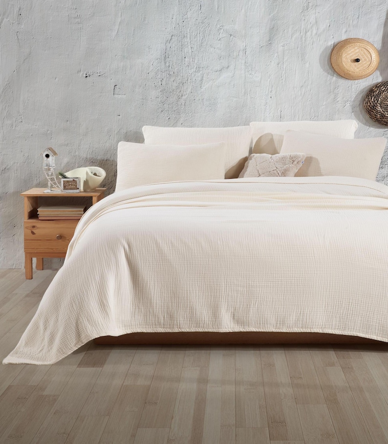 May include: A white waffle-weave throw blanket on a bed with white pillows. The bed is in a room with a white wall and a wooden bedside table.