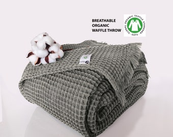 May include: Dark gray waffle throw blanket made from breathable organic cotton. The blanket has a waffle texture and a fringe border. The Global Organic Textile Standard (GOTS) certification label is visible on the blanket.