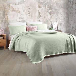 May include: A light green throw blanket with a fringed edge. The blanket is folded on a bed with white and pink pillows.