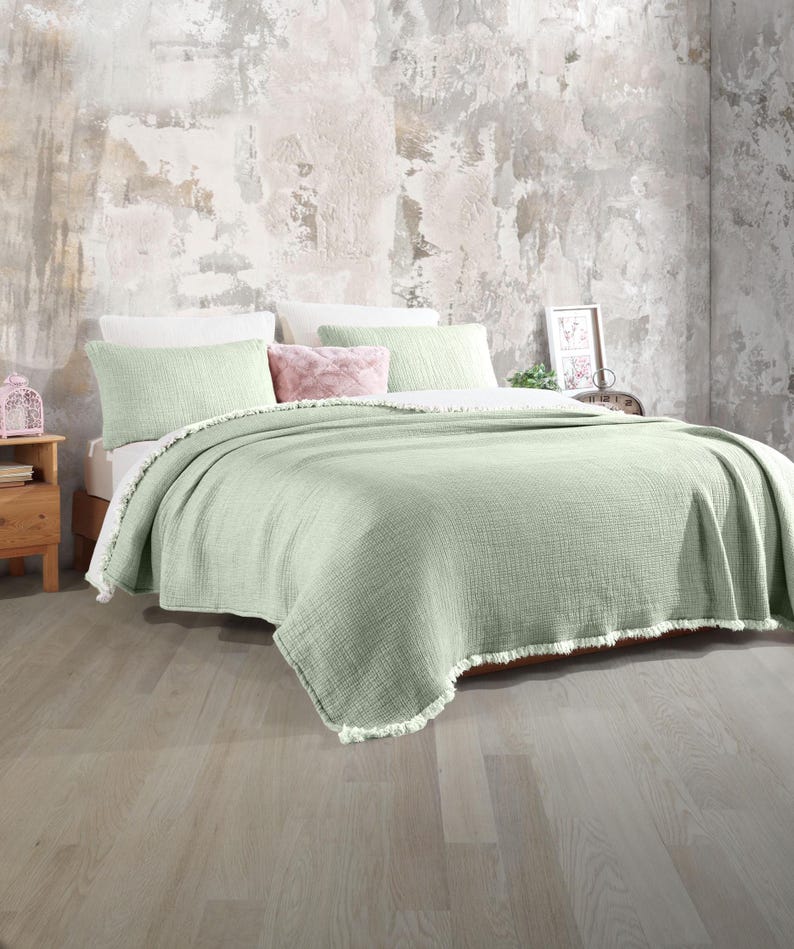 May include: A light green throw blanket with a fringed edge. The blanket is folded on a bed with white and pink pillows.