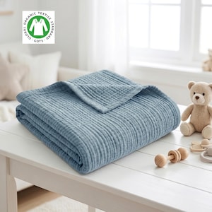 May include: A folded blue muslin blanket with a textured, multi-layered fabric surface. The blanket features a Global Organic Textile Standard GOTS logo in the corner. The fabric has a visible crinkled weave pattern throughout the entire material.