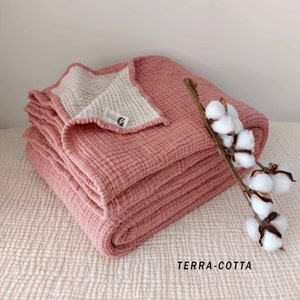 May include: Terracotta waffle weave throw blanket folded on a white textured surface. The blanket has a white cotton lining and a small label with the word "Organic" on it. A sprig of cotton is resting on the blanket.