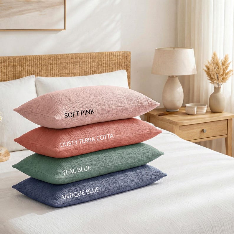 Pode incluir: Quatro almofadas decorativas retangulares em rosa suave, terracota empoeirada, azul-petr&oacute;leo e azul antigo. Cada almofada tem uma superf&iacute;cie texturizada e &eacute; rotulada com o nome da sua cor. As almofadas est&atilde;o empilhadas numa cama branca.
