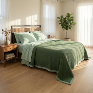 May include: A bed with a wooden headboard and a green waffle-weave blanket. The bed has light green sheets and pillows. A wooden nightstand with a vase of dried flowers and a stack of books sits beside the bed. The room has hardwood floors and sheer white curtains.
