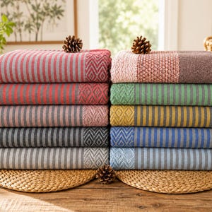 May include: A stack of ten rectangular woven cotton throws arranged in two columns. Each throw features a striped pattern with a diamond-textured border and fringed edges. The colors include red, pink, green, yellow, blue, gray, and brown tones.