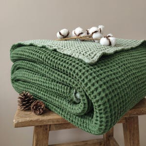 May include: A folded green waffle-weave blanket with a lighter green top layer. The blanket is stacked on a wooden stool, with pinecones and cotton sprigs as decoration. The blanket's texture is visible.