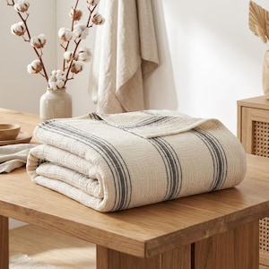 Könnte beinhalten: Eine gefaltete, cremefarbene Decke mit dunkelgrauen vertikalen Streifen. Die Decke besteht aus einem strukturierten Stoff und liegt auf einem Holztisch. Eine Vase mit Baumwollzweigen und eine Holzschale sind ebenfalls zu sehen.