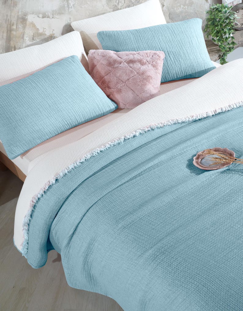 May include: A light blue throw blanket with a white fringe border. The blanket is folded on a bed with white and light blue pillows.