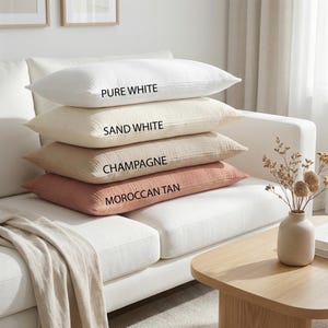 Pode incluir: Quatro almofadas retangulares em tons de branco e bege empilhadas em um sof&aacute; branco. A almofada superior &eacute; branco puro, seguida de branco areia, champanhe e marrom marroquino. Cada almofada tem uma superf&iacute;cie texturizada, com os nomes das cores.