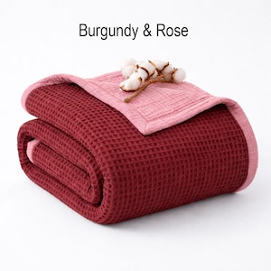 May include: A folded burgundy waffle-weave blanket with a rose-colored border and top layer. The text "Burgundy & Rose" is displayed at the top. A sprig of cotton adds a decorative touch to the blanket.