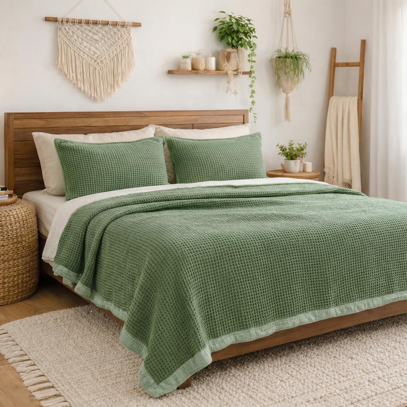 May include: A bed with a wooden headboard is covered with a green waffle-weave blanket and matching pillowcases. The bed is made up with a white sheet. A woven wall hanging and plants decorate the room, creating a cozy, inviting atmosphere.