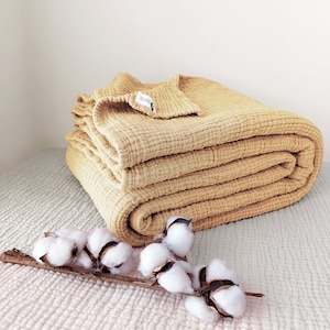May include: A mustard yellow waffle weave throw blanket folded on a light beige surface with a sprig of cotton bolls.
