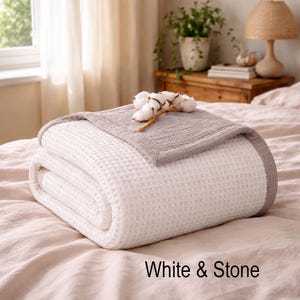 May include: A folded white and gray waffle-weave blanket with a cotton branch on top. The blanket is on a bed with a light pink sheet. The text "White & Stone" is at the bottom.