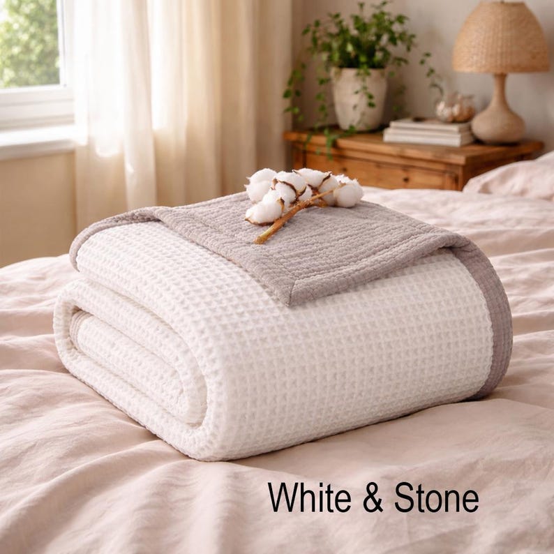 May include: A folded white and gray waffle-weave blanket with a cotton branch on top. The blanket is on a bed with a light pink sheet. The text "White & Stone" is at the bottom.