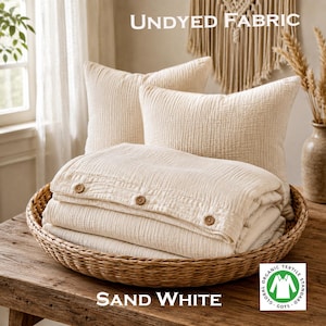 May include: Two square pillows and a folded duvet cover made of undyed fabric in a sand white color. The duvet cover features three visible brown buttons along the edge. The text UNDYED FABRIC and SAND WHITE appear, along with a GOTS certification logo.