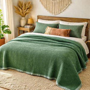May include: A bed is made with a green waffle-weave blanket and matching green pillows. A decorative fringed pillow in shades of orange and tan adds a contrasting accent. The bed is on a woven rug, and the headboard is made of wood.