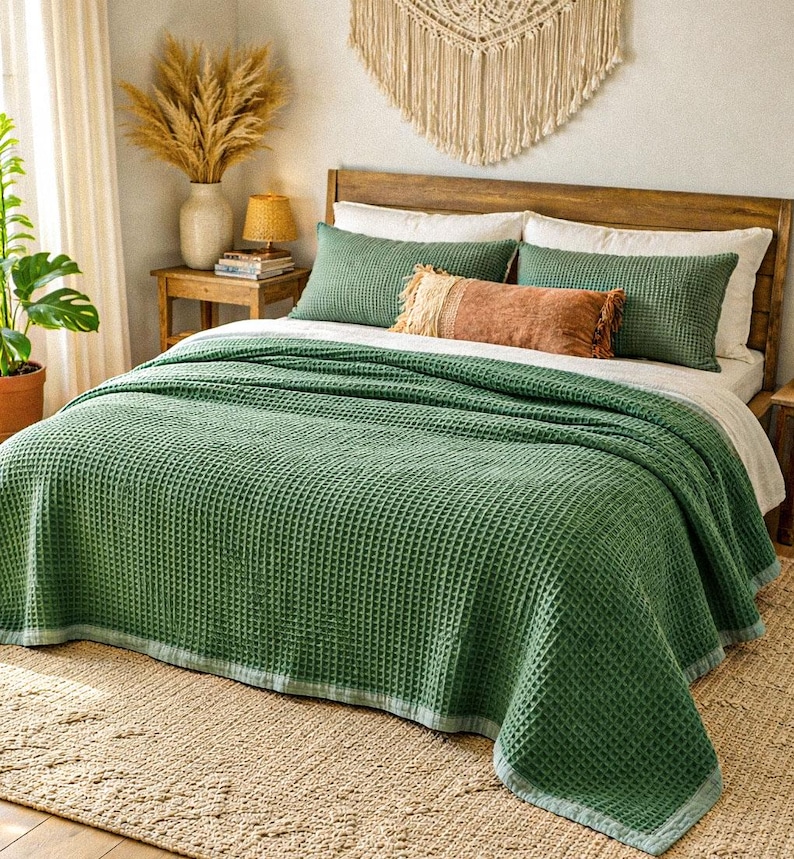 May include: A bed is made with a green waffle-weave blanket and matching green pillows. A decorative fringed pillow in shades of orange and tan adds a contrasting accent. The bed is on a woven rug, and the headboard is made of wood.