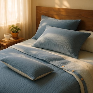 Pode incluir: Uma cama com roupa de cama azul, incluindo duas almofadas grandes, uma almofada longa e um cobertor dobrado. A roupa de cama tem uma apar&ecirc;ncia texturizada. Uma cabeceira de madeira e uma pequena mesa lateral com uma planta e uma x&iacute;cara tamb&eacute;m s&atilde;o vis&iacute;veis.