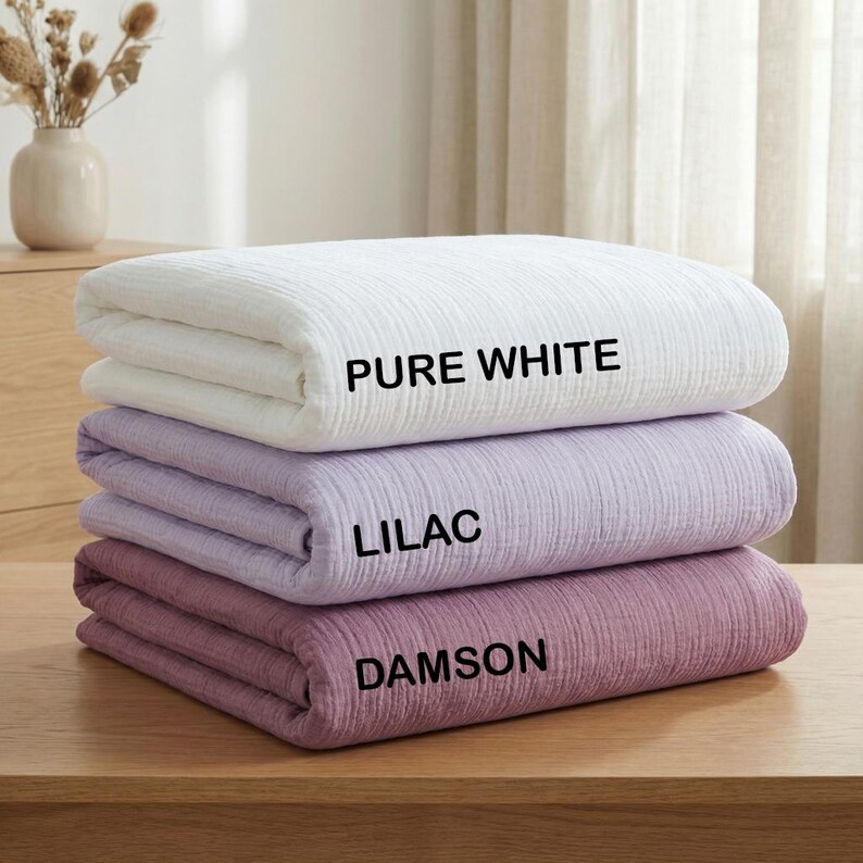 K&ouml;nnte beinhalten: Ein Stapel aus drei gefalteten Decken mit einer strukturierten, geknitterten Oberfl&auml;che. Die oberste Decke ist mit PURE WHITE beschriftet, die mittlere mit LILAC und die unterste mit DAMSON. Jede Decke ist ordentlich rechteckig zusammengelegt.