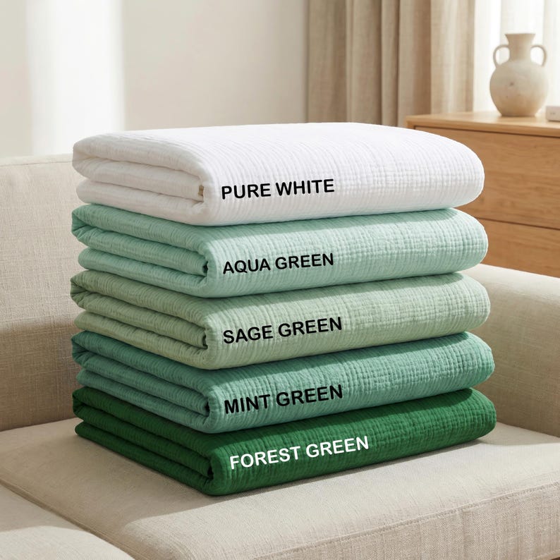 May include: A stack of five folded muslin blankets in varying shades of green and white. The colors are labeled as Pure White, Aqua Green, Sage Green, Mint Green, and Forest Green. Each blanket features a soft, crinkled texture and layered fabric.