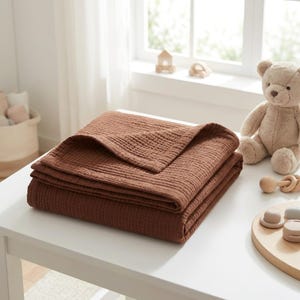 May include: A folded, solid rust-brown muslin blanket with a visible crinkled, multi-layered texture. The fabric features a square-stitched hem along the edges, showcasing a soft, lightweight appearance suitable for layering or covering.