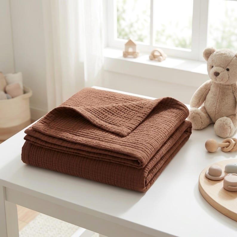 May include: A folded, solid rust-brown muslin blanket with a visible crinkled, multi-layered texture. The fabric features a square-stitched hem along the edges, showcasing a soft, lightweight appearance suitable for layering or covering.