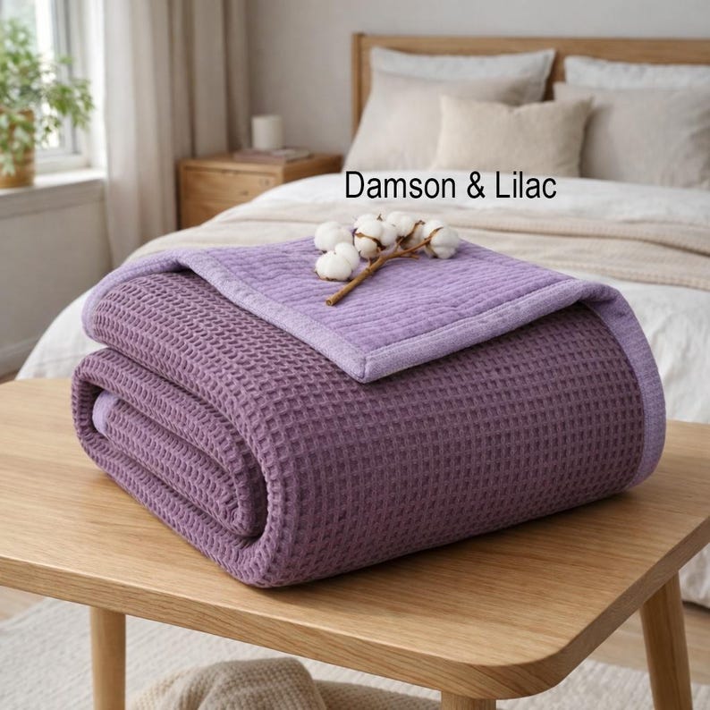 May include: A folded, damson and lilac colored waffle-weave blanket rests on a wooden table. The blanket is topped with a lighter lilac section and a sprig of cotton. The text "Damson & Lilac" is visible.