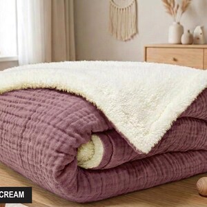 K&ouml;nnte beinhalten: Eine gefaltete Decke in Pflaume und Creme. Die Decke hat eine strukturierte, gesteppte Optik auf der Pflaumenseite und eine weiche, cremefarbene, flauschige Textur auf der R&uuml;ckseite. Der Text "DAMSON & CREAM" befindet sich in einem schwarzen Feld.