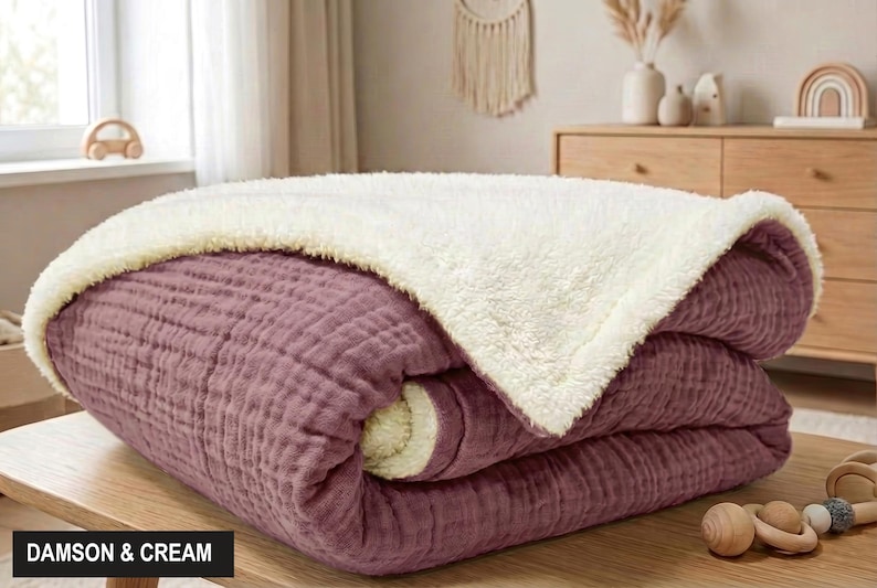 K&ouml;nnte beinhalten: Eine gefaltete Decke in Pflaume und Creme. Die Decke hat eine strukturierte, gesteppte Optik auf der Pflaumenseite und eine weiche, cremefarbene, flauschige Textur auf der R&uuml;ckseite. Der Text "DAMSON & CREAM" befindet sich in einem schwarzen Feld.