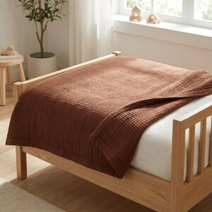 May include: A solid rust-colored, textured muslin blanket draped over a light-toned wooden bed frame. The blanket features a visible grid-like weave pattern and is layered over a white fitted sheet on a bed with vertical wooden slats.