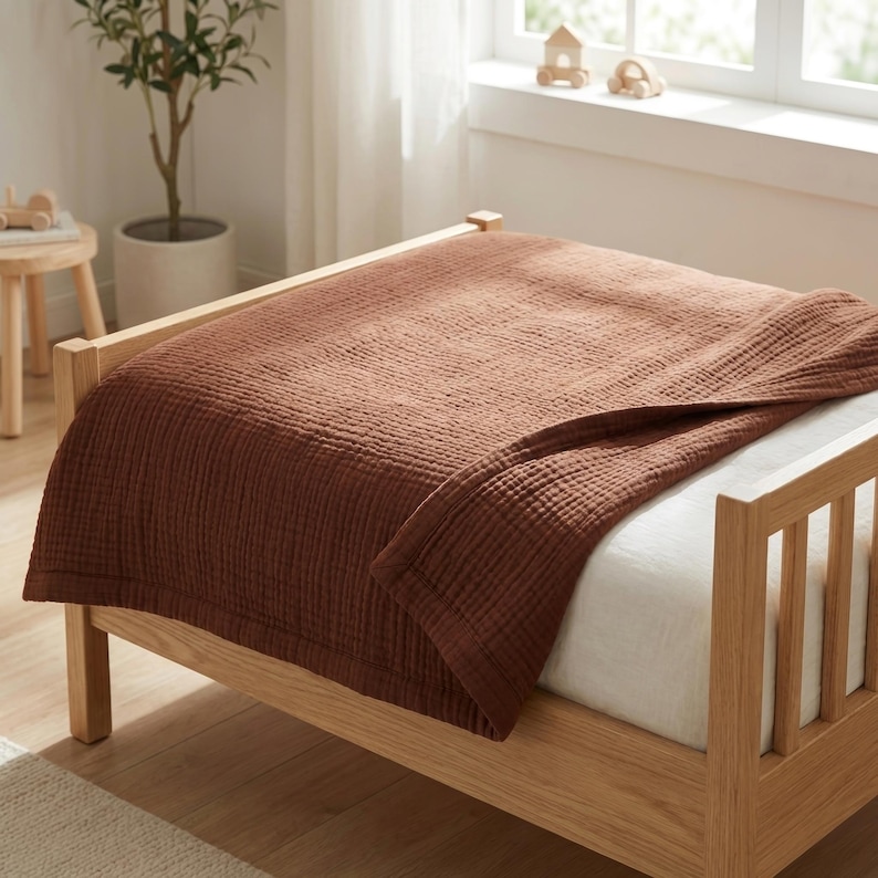 May include: A solid rust-colored, textured muslin blanket draped over a light-toned wooden bed frame. The blanket features a visible grid-like weave pattern and is layered over a white fitted sheet on a bed with vertical wooden slats.