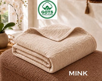 May include: A folded light beige blanket with a textured, crinkled surface. The blanket features a GOTS Certified Textiles logo and the word MINK printed in white. The blanket is resting on a brown, textured, fuzzy surface.