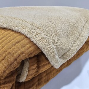 K&ouml;nnte beinhalten: Eine gefaltete Decke mit einem strukturierten, gerippten, senffarbenen Stoff auf der Unterseite und einer weichen, cremefarbenen, flauschigen oberen Schicht. Die Decke scheint aus zwei verschiedenen Materialien zu bestehen.