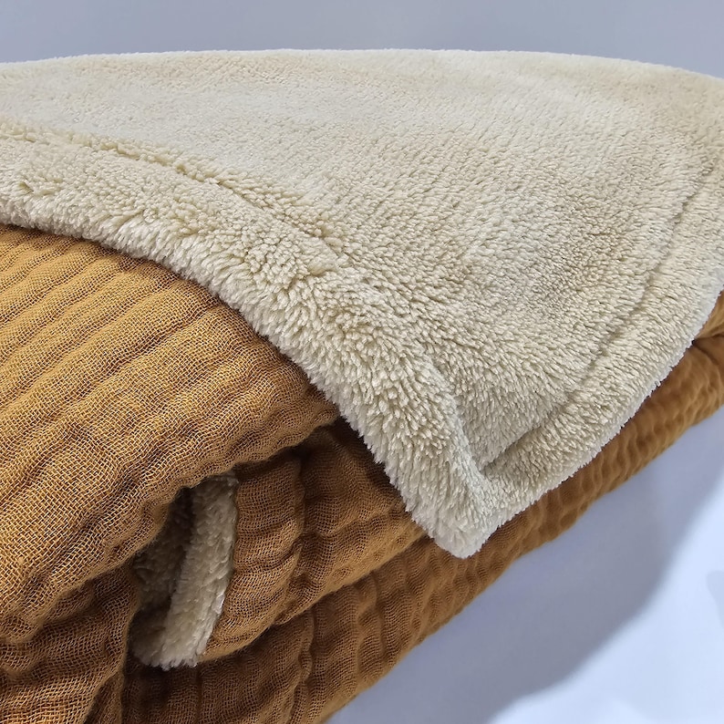 K&ouml;nnte beinhalten: Eine gefaltete Decke mit einem strukturierten, gerippten, senffarbenen Stoff auf der Unterseite und einer weichen, cremefarbenen, flauschigen oberen Schicht. Die Decke scheint aus zwei verschiedenen Materialien zu bestehen.