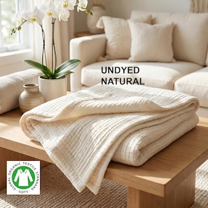 Op de afbeelding: Een gevouwen, ongeverfde, natuurkleurige katoenen deken met een getextureerd, gewatteerd oppervlak. De tekst "UNDYED NATURAL" is zichtbaar boven de deken en een groen-wit GOTS Global Organic Textile Standard-logo staat in de hoek.