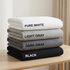 May include: A stack of four folded blankets with a textured, crinkled fabric surface. The blankets are labeled with black text as PURE WHITE, LIGHT GRAY, DARK GRAY, and BLACK, arranged from the top to the bottom of the stack in that order.