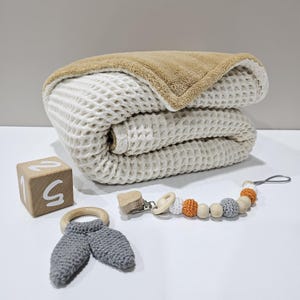 May include: A folded beige waffle-knit baby blanket with a tan plush backing. A wooden block with the number 5, a grey crochet bunny ear teether, and a beaded dummy clip with wooden and crochet beads are also present.