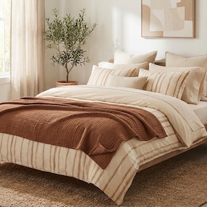 May include: A bed featuring a beige and white striped duvet cover, multiple matching pillows, and a textured rust-colored waffle-weave throw blanket draped across the foot. The bed frame is made of wood and sits on a woven jute area rug.