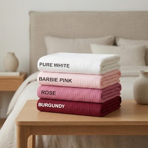 May include: A stack of four folded textured blankets in a gradient of colors. The top blanket is labeled PURE WHITE, the second is BARBIE PINK, the third is ROSE, and the bottom blanket is labeled BURGUNDY. Each blanket features a ribbed fabric.