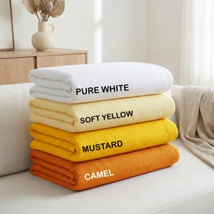 May include: A stack of four folded muslin blankets in a textured fabric. The colors from top to bottom are labeled as PURE WHITE, SOFT YELLOW, MUSTARD, and CAMEL. Each blanket is neatly layered on top of the other, showcasing a consistent weave.