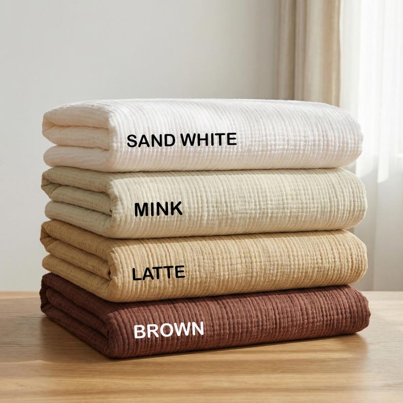 May include: A stack of four folded rectangular blankets with a textured, crinkled fabric surface. The blankets are labeled with text: SAND WHITE for the top, MINK for the second, LATTE for the third, and BROWN for the bottom blanket.