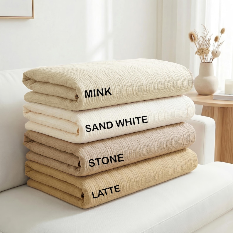 May include: A stack of four folded textured blankets in neutral tones. The blankets are labeled with black text as Mink, Sand White, Stone, and Latte, representing the color variations of the fabric layers stacked neatly on top of each other.