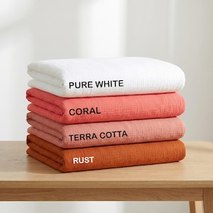 May include: A stack of four folded muslin blankets in solid colors. The top blanket is labeled PURE WHITE, the second is labeled CORAL, the third is labeled TERRA COTTA, and the bottom blanket is labeled RUST. Each blanket has a textured fabric.