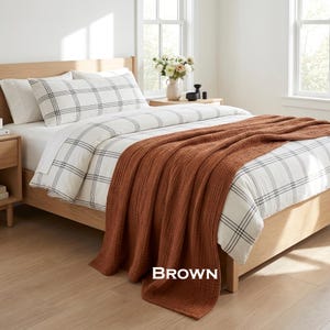 K&ouml;nnte beinhalten: Ein Bett mit einem hellen Holzrahmen, wei&szlig;er Bettw&auml;sche mit schwarzem Karomuster und einer strukturierten, einfarbig braunen Tagesdecke, die &uuml;ber das Fu&szlig;ende drapiert ist. Das Wort BROWN steht in wei&szlig;er Schrift unten in der Bildmitte.